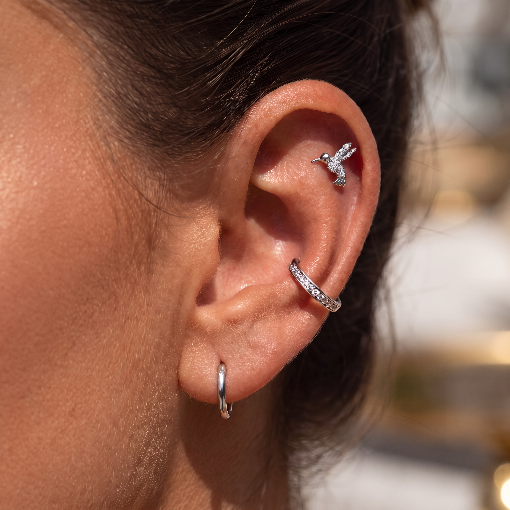 Close-up of a person's ear showcasing a multi-piece silver ear cuff set, including a small hummingbird stud, a curved ear cuff with clear stones, and a simple hoop earring.