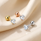 Three heart-shaped crystal labret stud earrings in gold, rose gold, and silver, arranged on a draped beige satin fabric background.