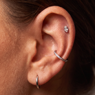 Close-up of a person's ear showcasing three silver-toned ear piercings: a small hoop in the lobe, a pavé-set huggie ring on the helix, and a small cluster stud on the lobe.