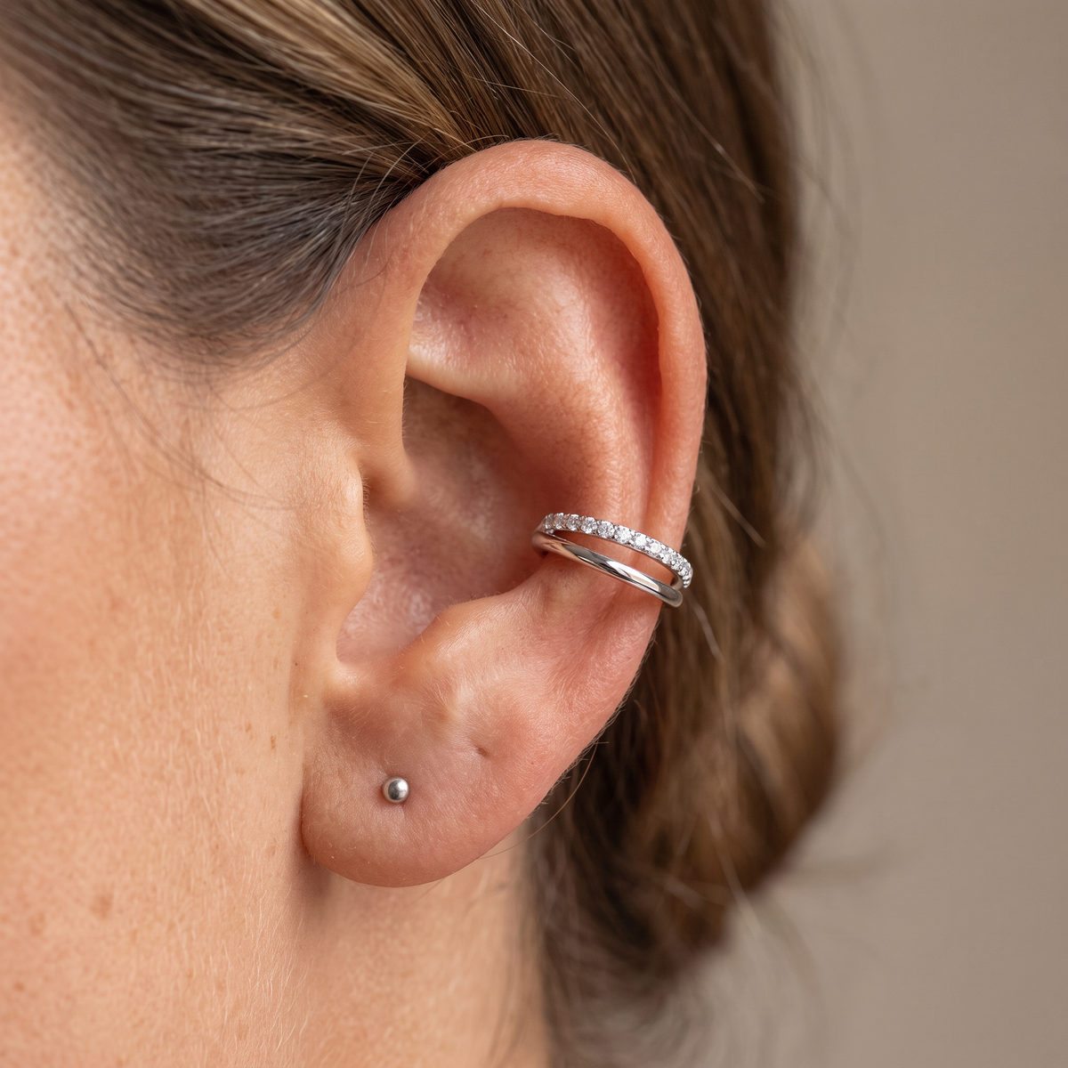 A close-up of a person's ear wearing a thin silver ear cuff adorned with small clear stones, along with a small stud earring, against a soft beige background.