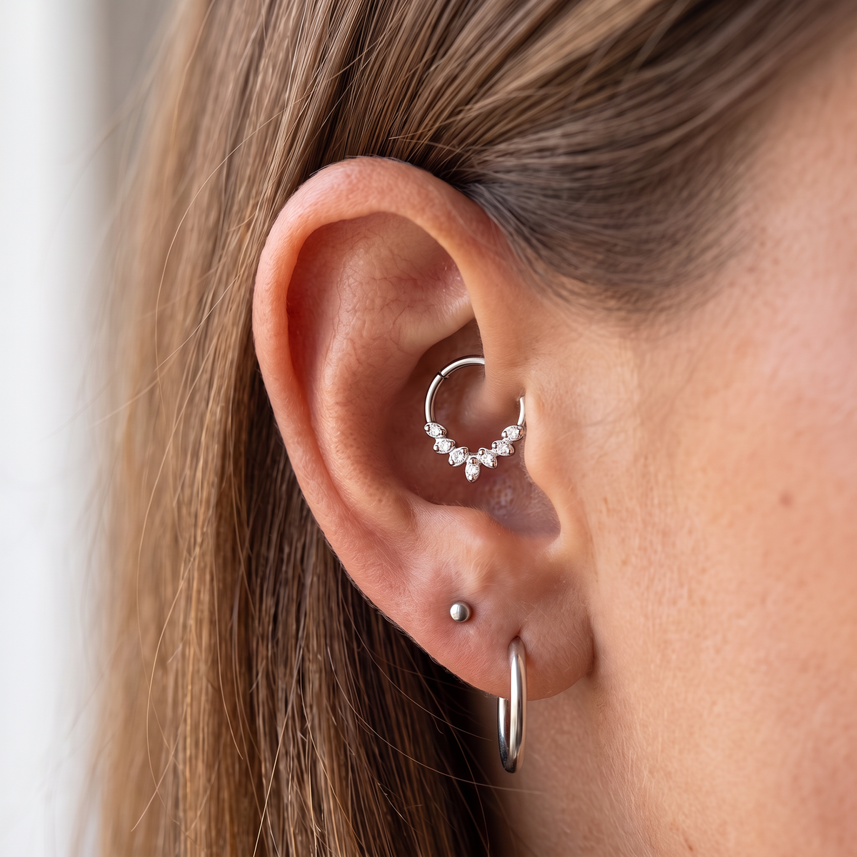 Close-up of a person's ear showcasing silver hoop and cartilage piercings, including a small cluster of clear stones on a hoop in the upper ear cartilage and a simple silver hoop in the lobe.