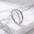 A silver double-band ring with one band paved with small clear stones, displayed on a white marble block.