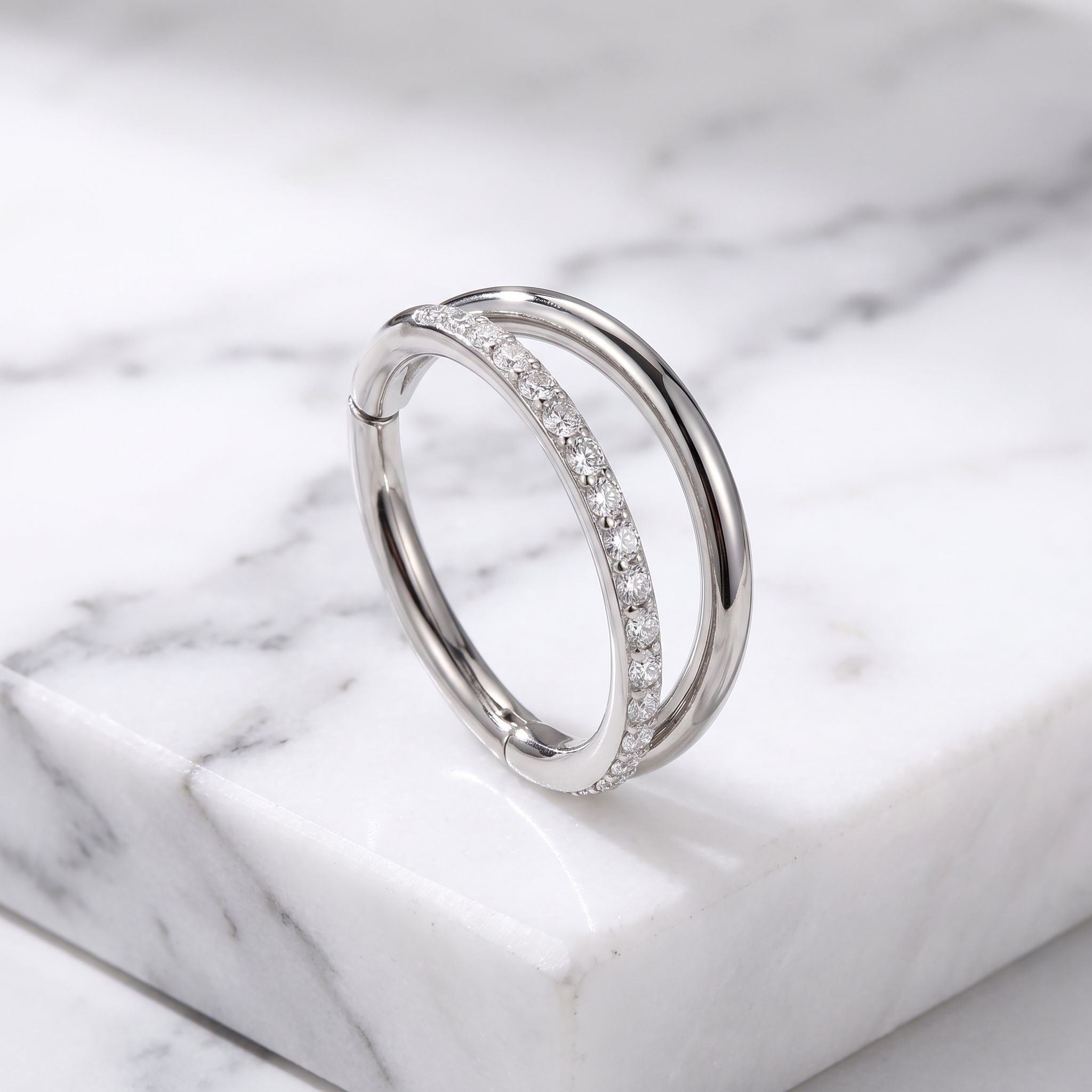 A silver double-band ring with one band paved with small clear stones, displayed on a white marble block.