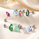 Six silver-toned barbell stud earrings with teardrop-shaped faceted gemstones in pink, iridescent blue/purple, iridescent green/blue, iridescent blue/purple, green, and pale blue, arranged on a beige satin fabric background.
