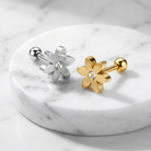 Two flower-shaped stud earrings displayed on a round white marble tray. One earring is silver-toned with a clear crystal center, and the other is gold-toned with a clear crystal center.