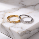 Two slim hoop rings with small clear stones, one in yellow gold and one in silver, displayed on a white marble slab.