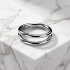 A polished silver-toned metal cuff bracelet with three parallel bands, resting on a white marble block with gray veining.