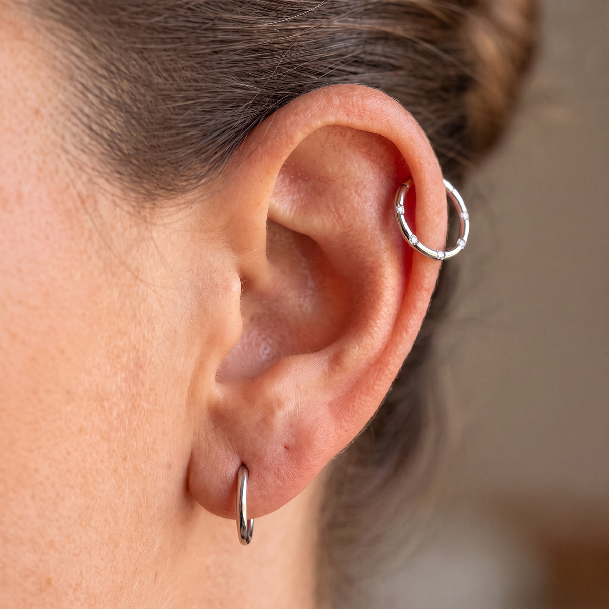 Close-up of a person's ear showing two silver hoop earrings, one on the lobe and one on the helix, with brown hair pulled back.