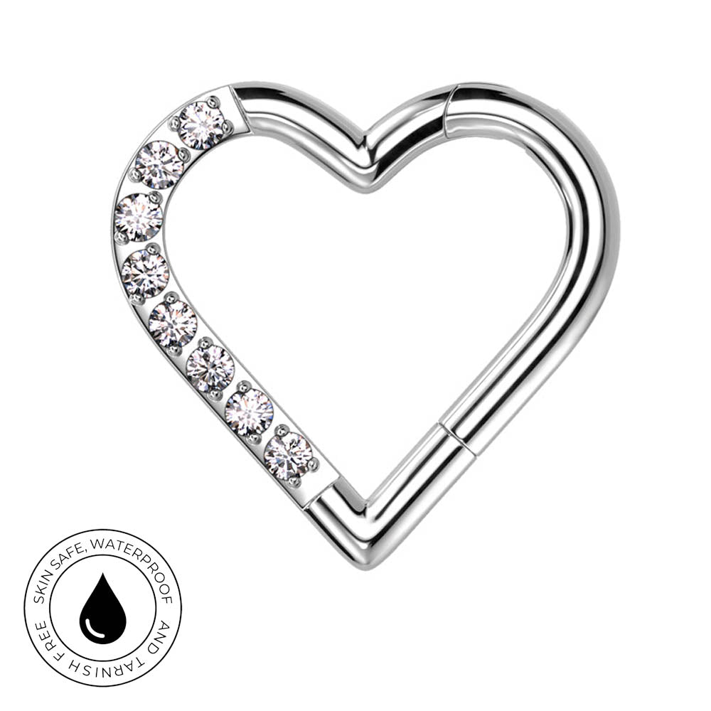 A silver-toned heart-shaped body jewelry clip with a row of clear rhinestones along one side, shown on a plain white background with a circular skin-safe waterproof badge in the lower-left corner.