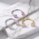 Three open hoop rings displayed on a white marble slab: two silver-toned rings with pink gemstones and one gold-toned ring with clear gemstones, photographed in a clean studio setting.