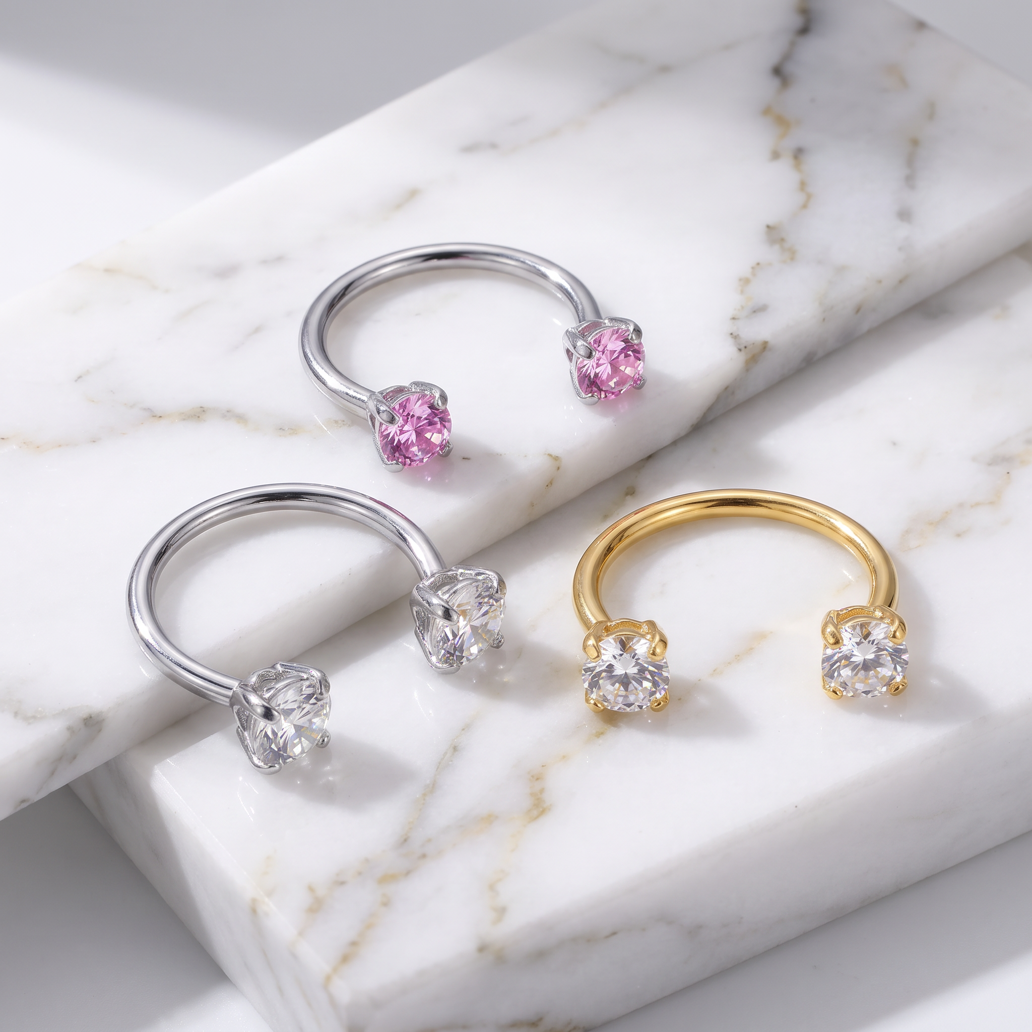 Three open hoop rings displayed on a white marble slab: two silver-toned rings with pink gemstones and one gold-toned ring with clear gemstones, photographed in a clean studio setting.