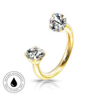 A gold-plated open ring piercing with two round-cut clear gemstones, shown on a white background with a small circular skin-safe waterproof badge in the lower-left corner.