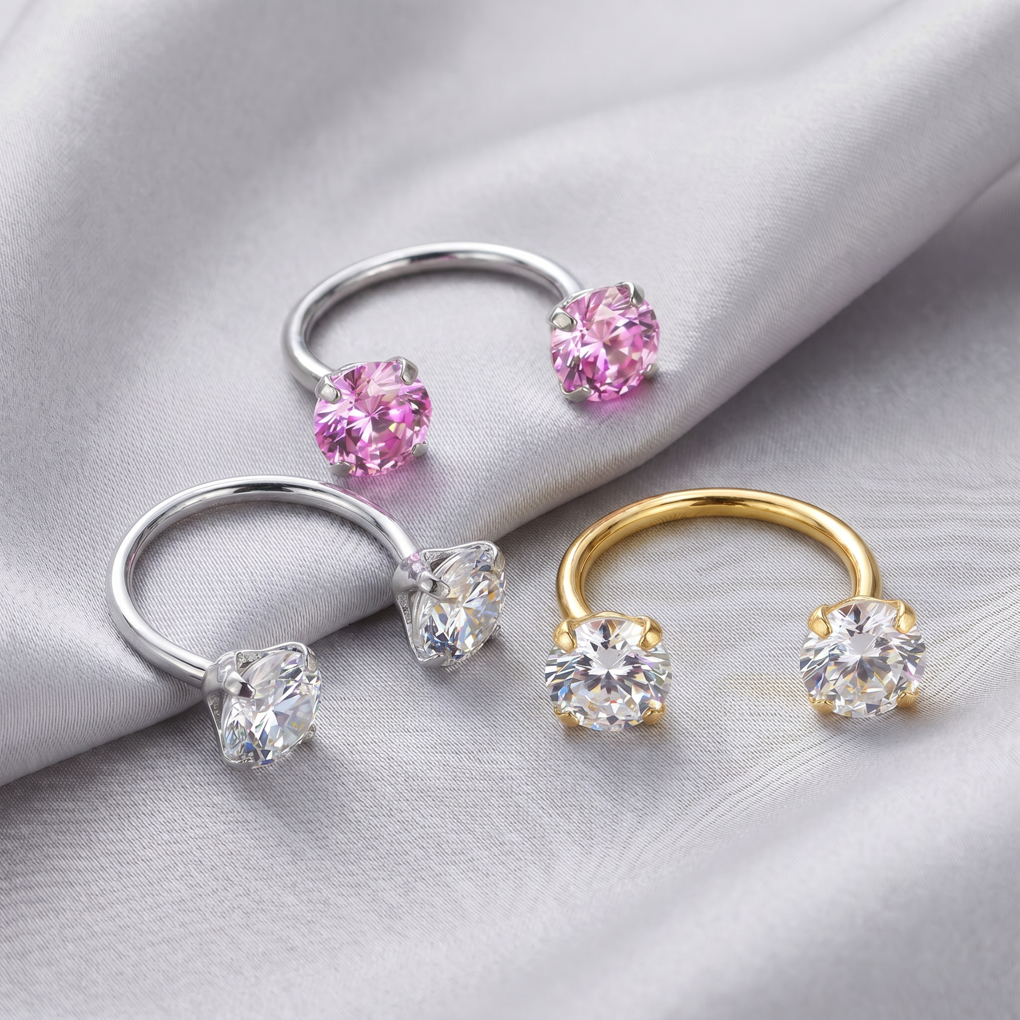 Three U-shaped ring ear cuffs displayed on draped satin fabric, featuring silver, yellow gold, and rose gold metal bands with clear gemstone settings and pink gemstone settings.