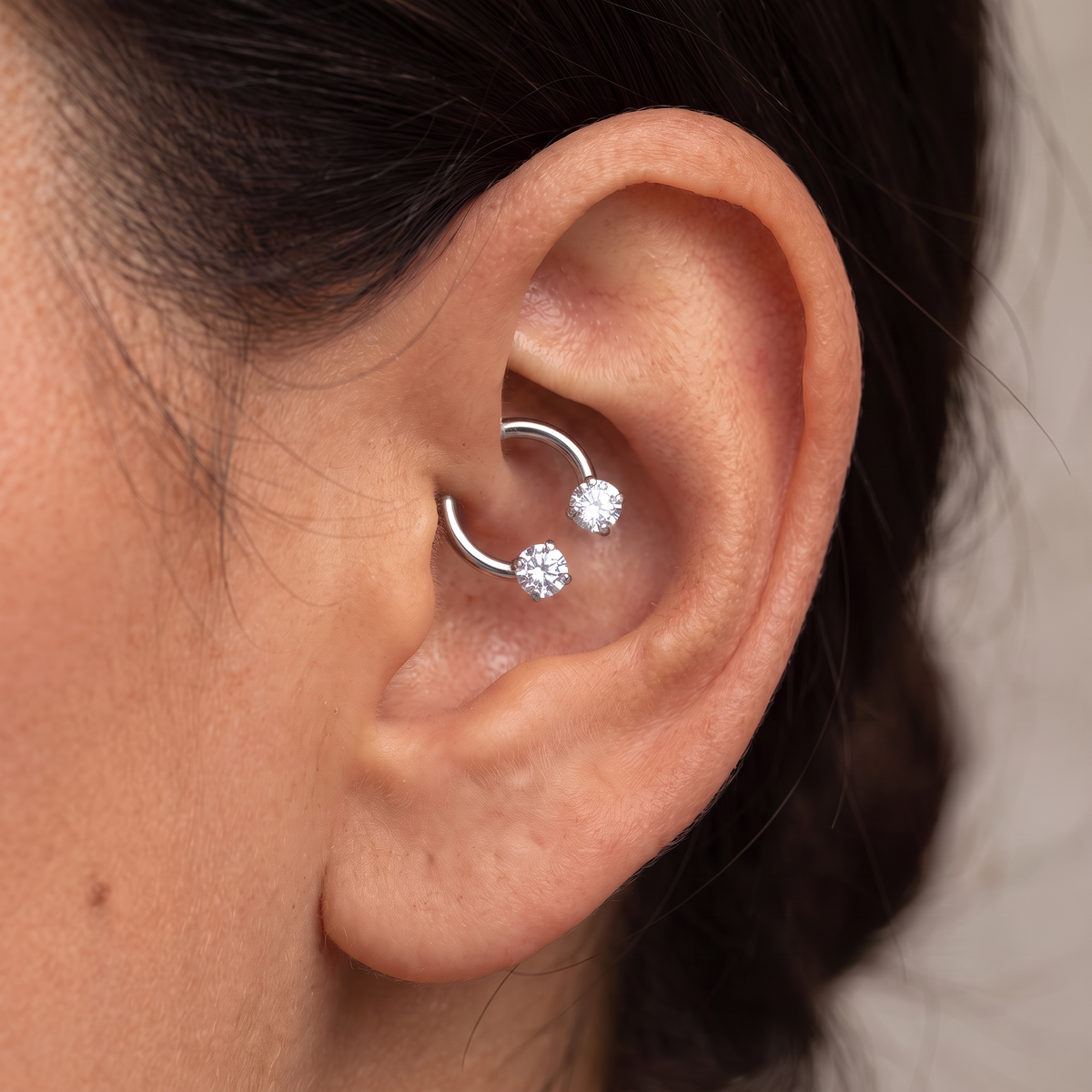 A close-up of a person's ear wearing a silver cartilage hoop piercing with two small clear gemstone studs.