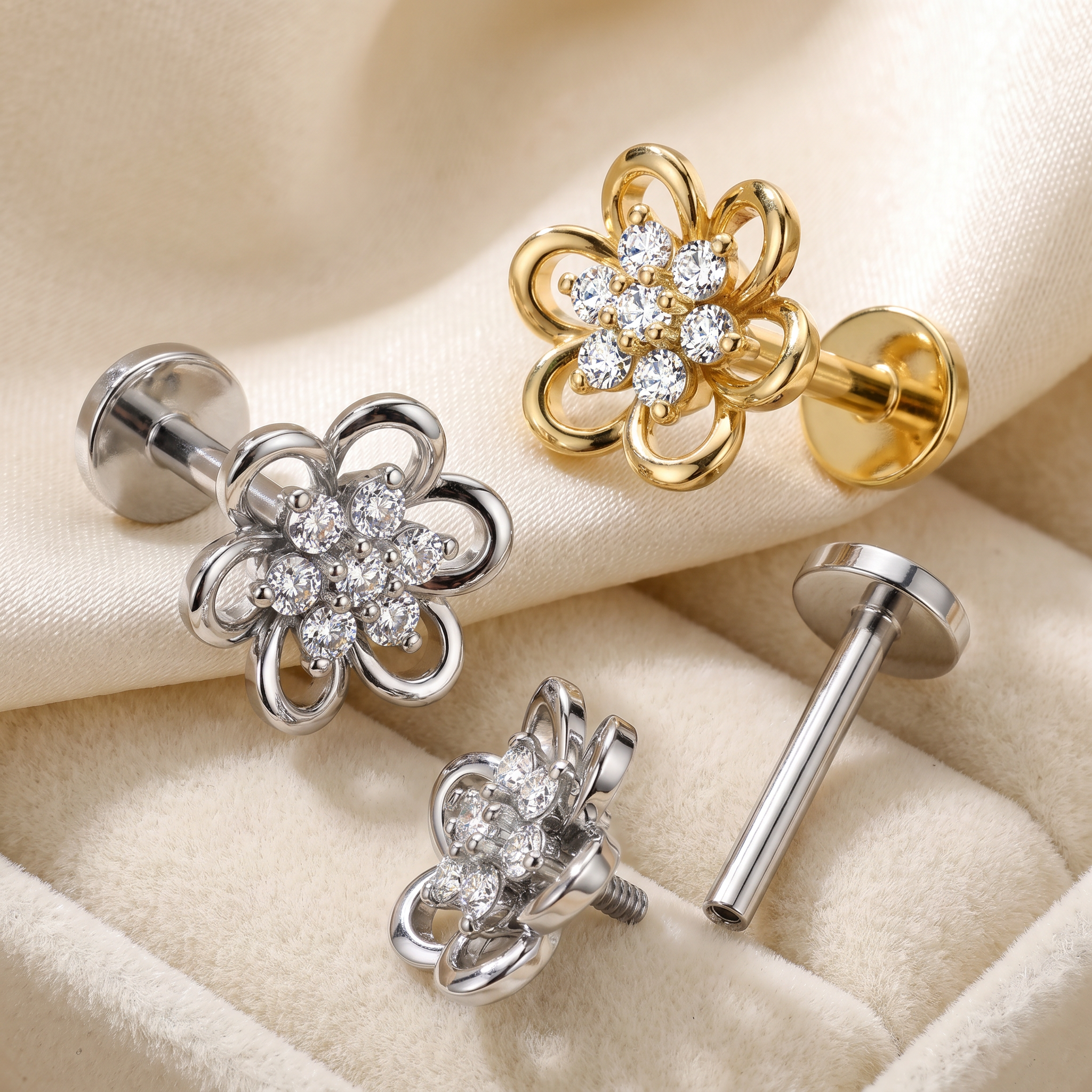 Four flower-shaped stud earrings displayed on a beige fabric surface, including two silver-toned and two gold-toned versions, each with clear rhinestone detailing and a straight post back.