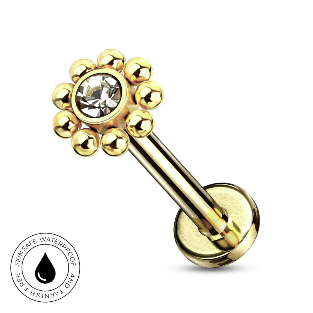 Gold-tone labret piercing barbell with a flower-shaped top featuring a clear faceted center stone and surrounding gold beads, shown on a white background with a circular skin-safe waterproof badge in the lower left.
