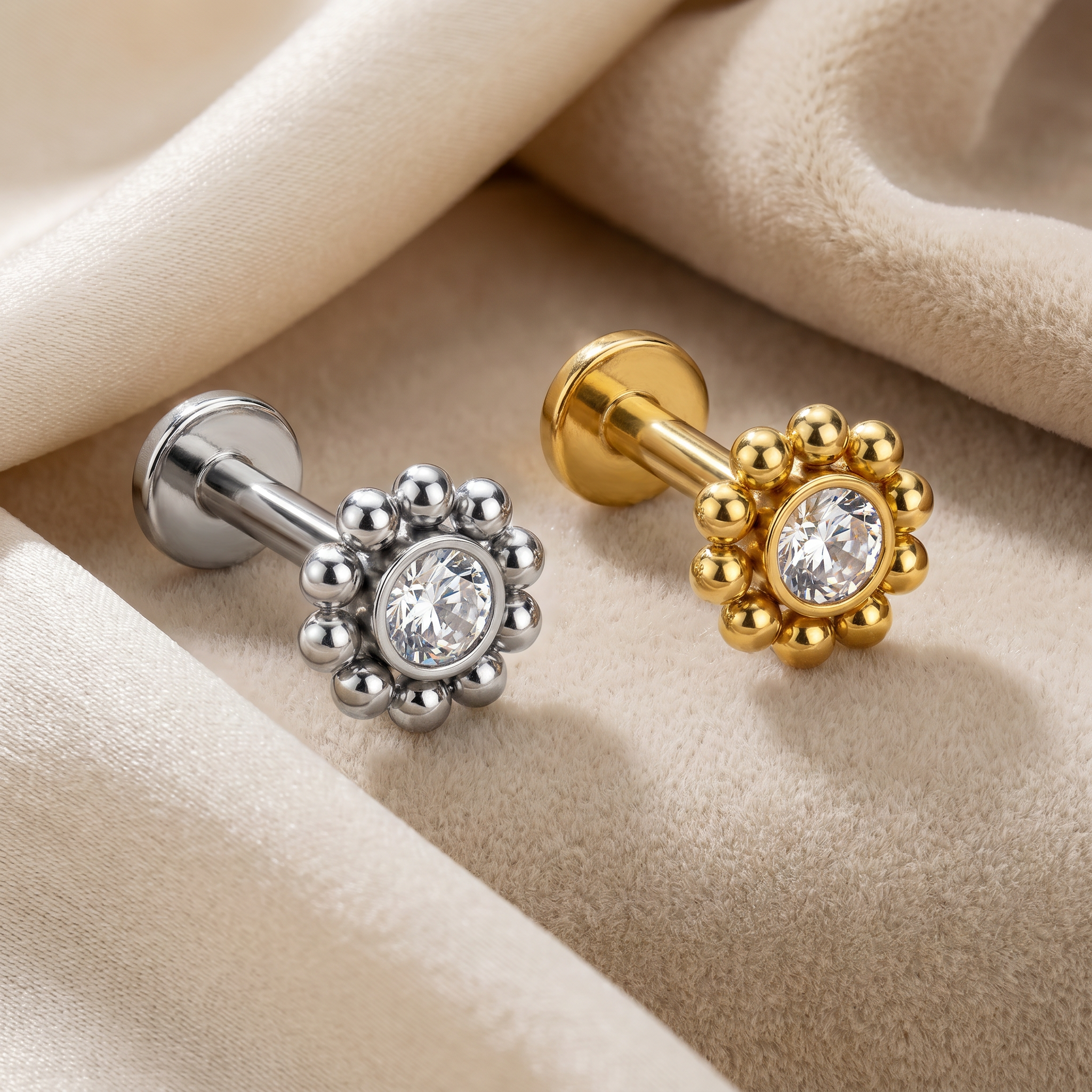 Two flower-shaped labret studs with clear round center stones, one in silver-tone metal and one in gold-tone metal, displayed on draped beige fabric.