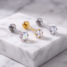Three barbell stud earrings displayed on a white marble block. One earring has a silver-toned post with a clear faceted gemstone, one has a gold-toned post with a clear faceted gemstone, and one has a silver-toned post with an iridescent clear gemstone.
