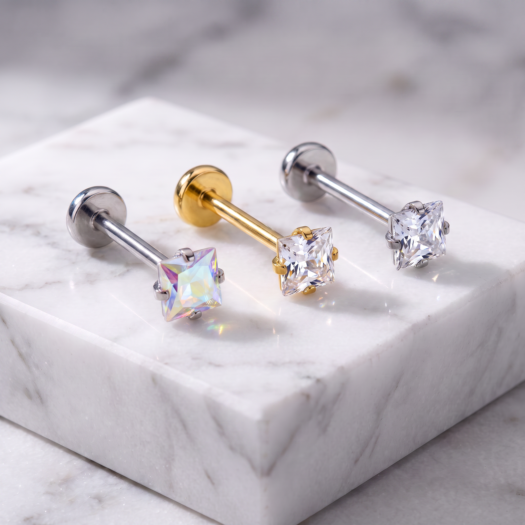 Three barbell stud earrings displayed on a white marble block. One earring has a silver-toned post with a clear faceted gemstone, one has a gold-toned post with a clear faceted gemstone, and one has a silver-toned post with an iridescent clear gemstone.