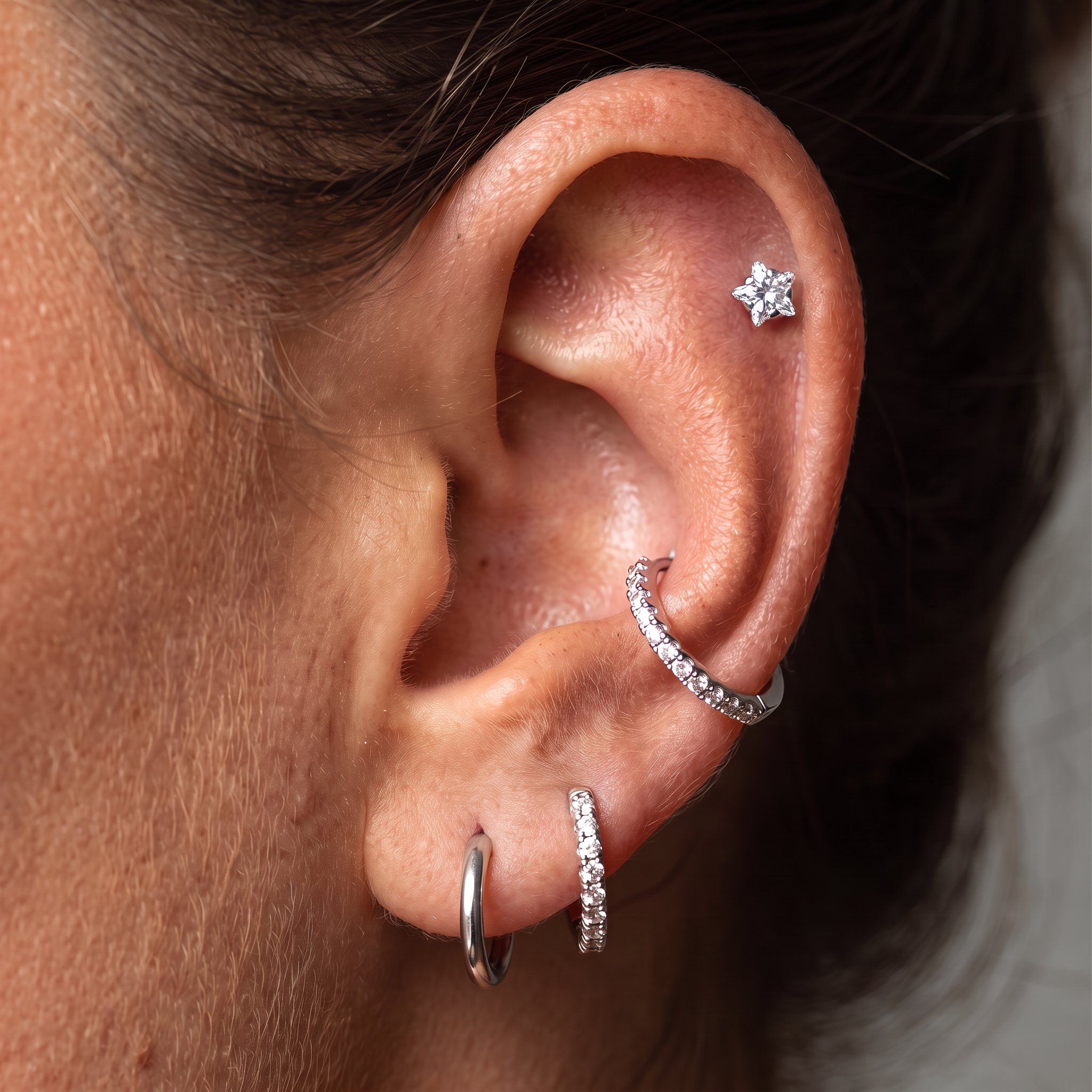 Close-up of a person's ear showcasing multiple silver-toned piercings: a small star-shaped stud in the lobe, a curved hoop with clear stones in the cartilage, a short vertical hoop with clear stones, and a simple curved hoop in the earlobe.
