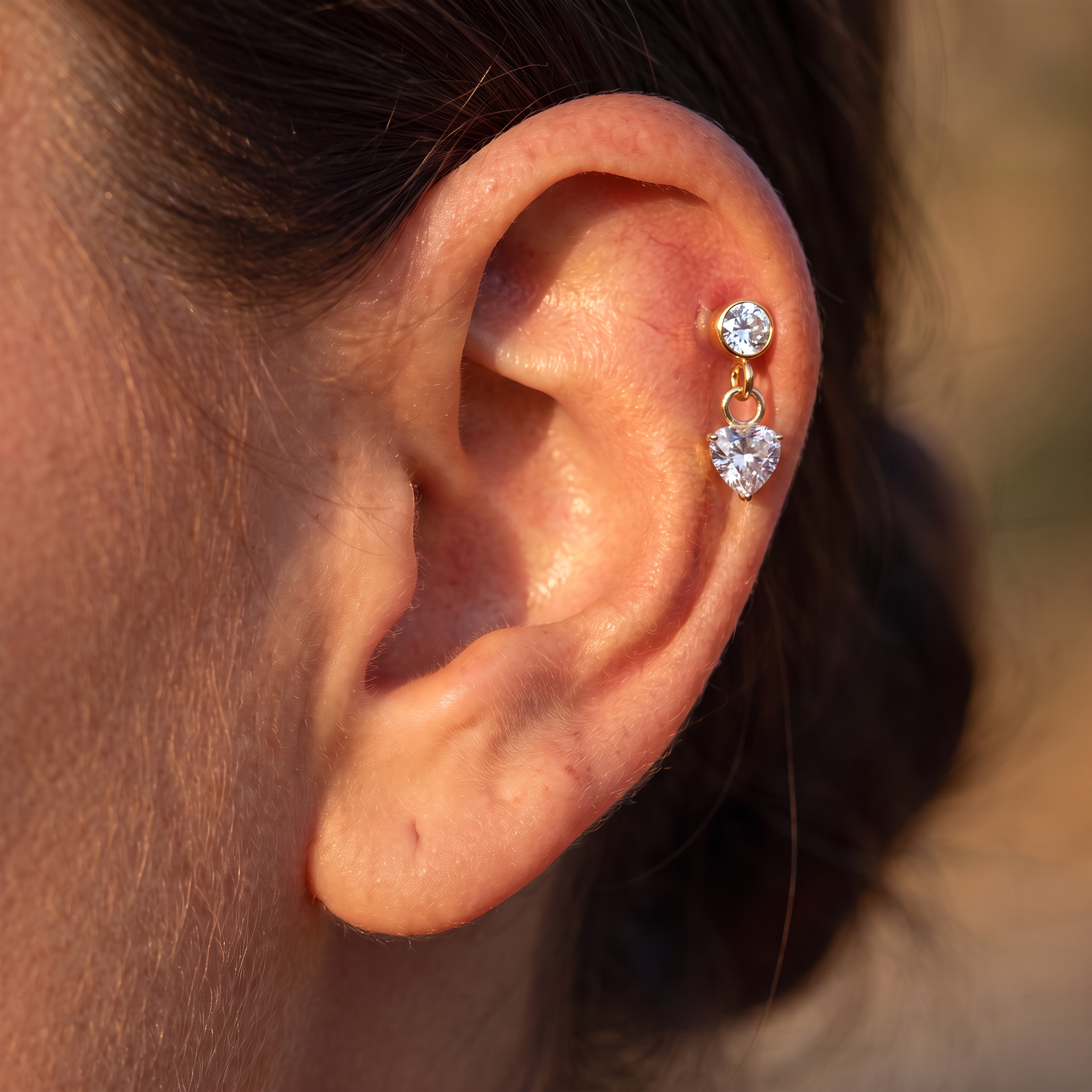 A close-up of a person's ear wearing a gold ear cuff with two dangling clear heart-shaped and round clear gemstones.