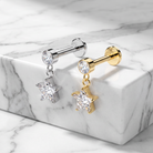 Two star-shaped dangle earrings displayed on a white marble block. One earring is silver-toned with a clear faceted gemstone, and the other is gold-toned with a matching clear gemstone.