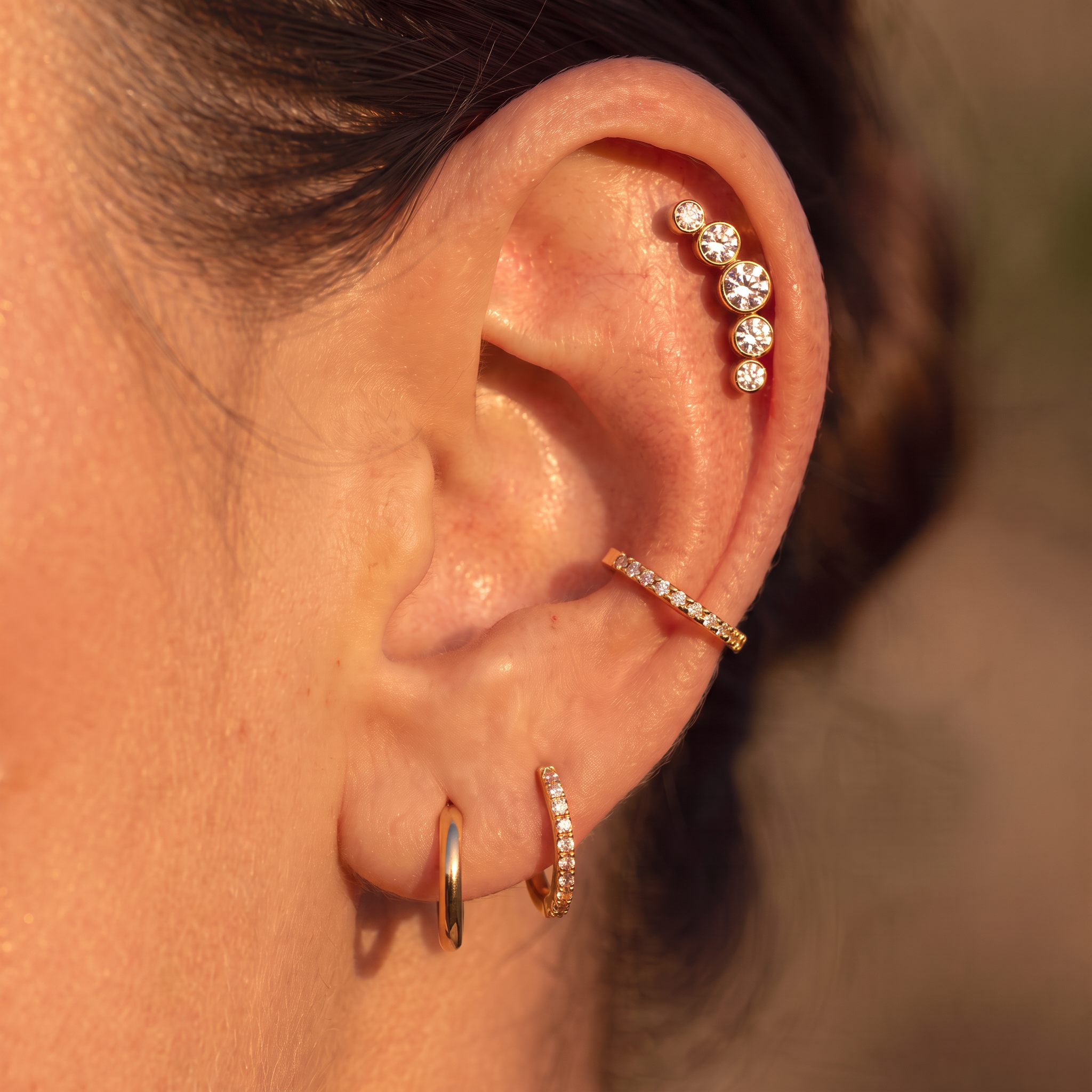 Close-up of a person's ear showcasing multiple gold ear piercings: a small hoop in the lobe, a curved hoop with tiny clear stones in the cartilage, and a curved barbell stud with clear stones on the upper ear.