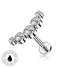 A silver-toned barbell-style body jewelry piece with four clear round gemstones, shown on a white background with a small circular skin-safe waterproof badge in the lower left.