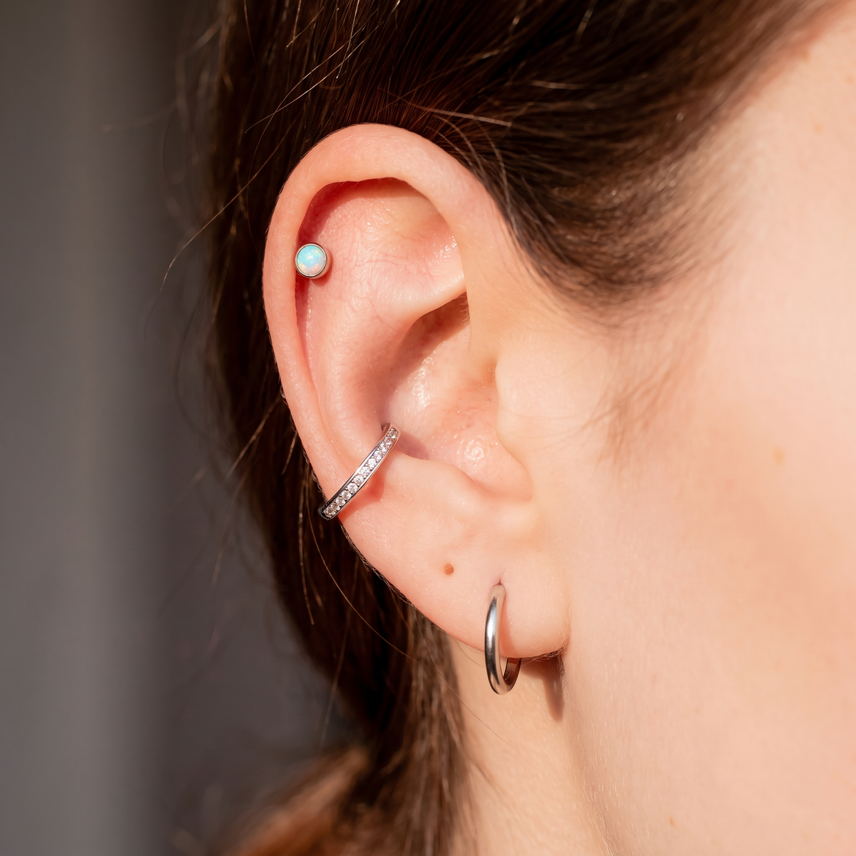 Close-up of a person's ear showcasing multiple piercings: a small round light blue stud in the lobe, a silver pavé hoop in the cartilage, and a silver hoop in the lobe.