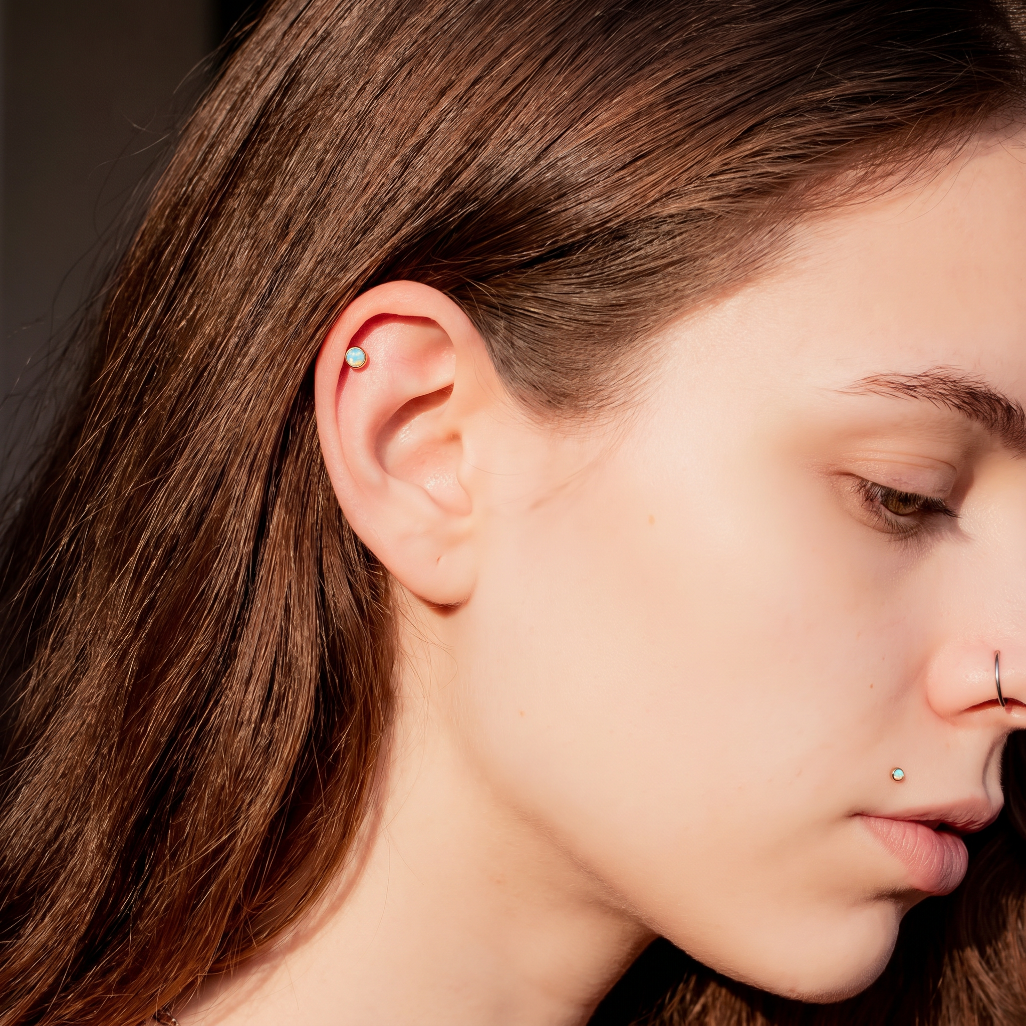Close-up of a person's ear wearing a small silver hoop earring, with brown hair, soft lighting, and a blurred dark background.