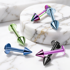 Four metallic curved barbell piercings in different colors are arranged on a white marble surface and a round marble pedestal. The colors visible are iridescent purple and blue, light blue, mint green, and black with a magenta-pink tip.