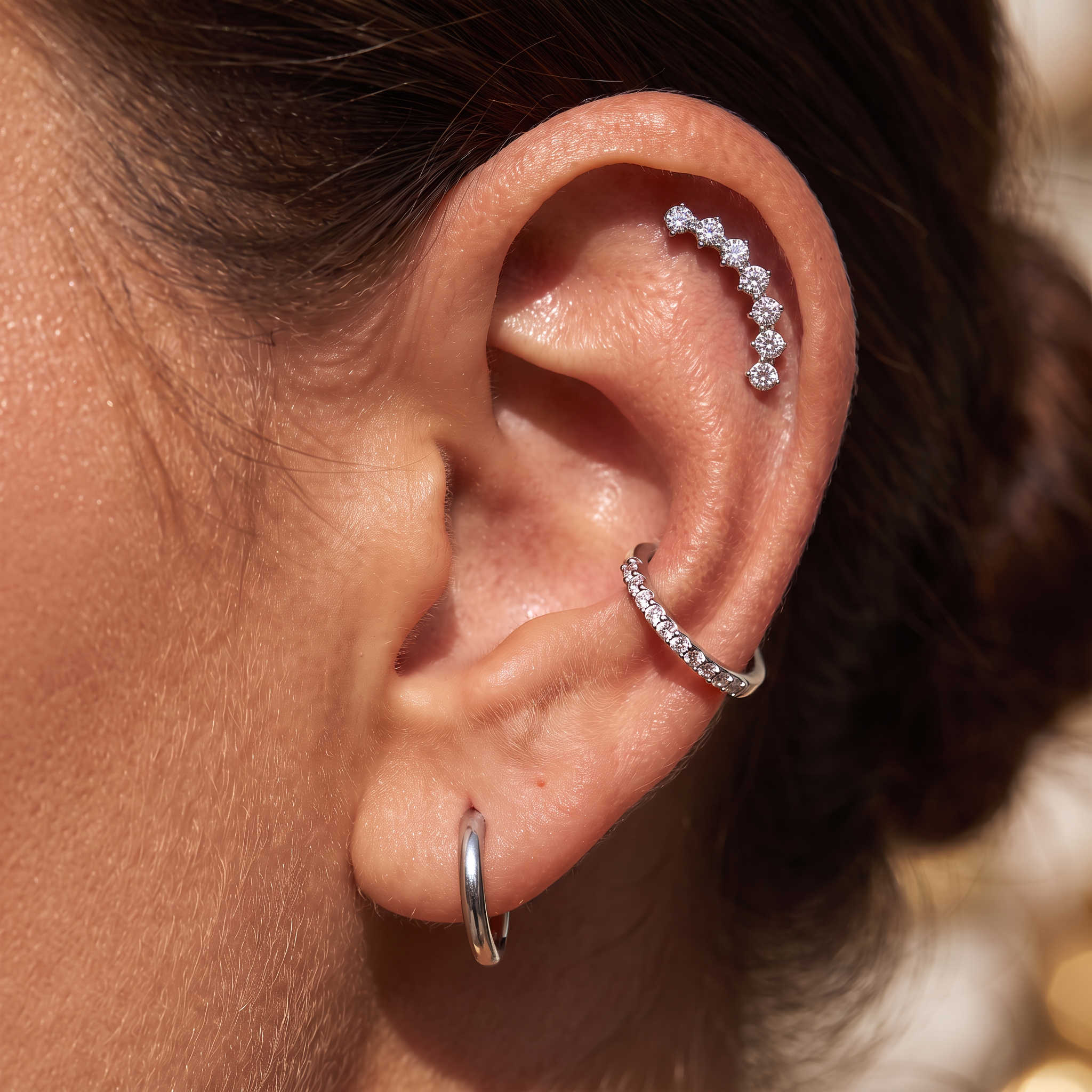 Close-up of a person's ear showcasing a multi-piece ear cuff stack: a small silver hoop in the lobe, a curved silver hoop set with clear stones across the helix, and a curved silver hoop set with clear stones along the upper ear cartilage.