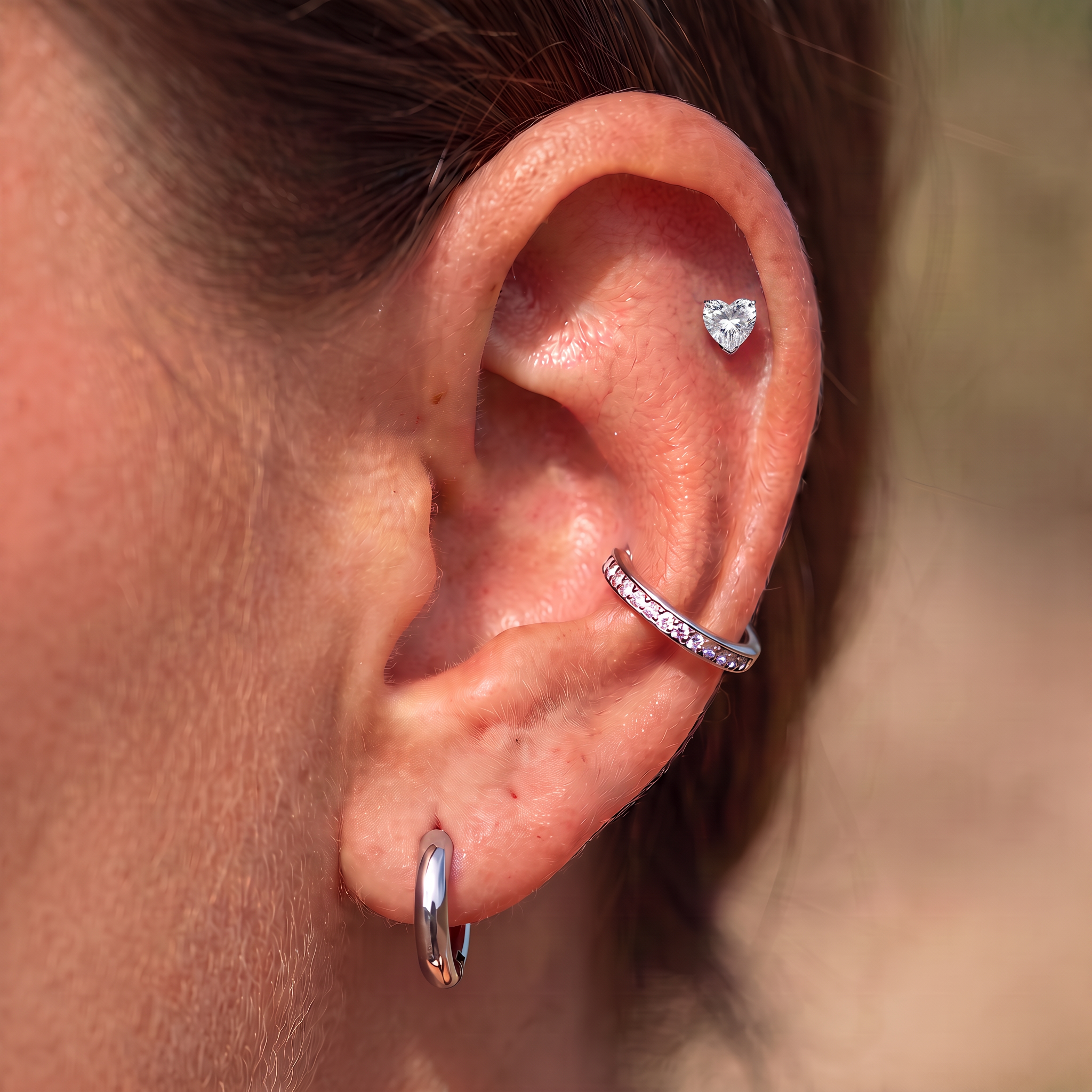 Close-up of a person's ear showcasing three silver ear piercings: a small star-shaped stud, a curved cartilage hoop set with clear stones, and a flat hoop earring.