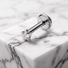 A silver-toned labret piercing stud with a clear gemstone, resting on a white marble block with gray veining.