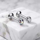 Two silver-toned barbell stud earrings displayed on a white marble block. One stud features a faceted clear crystal, while the other features a round iridescent rainbow crystal.