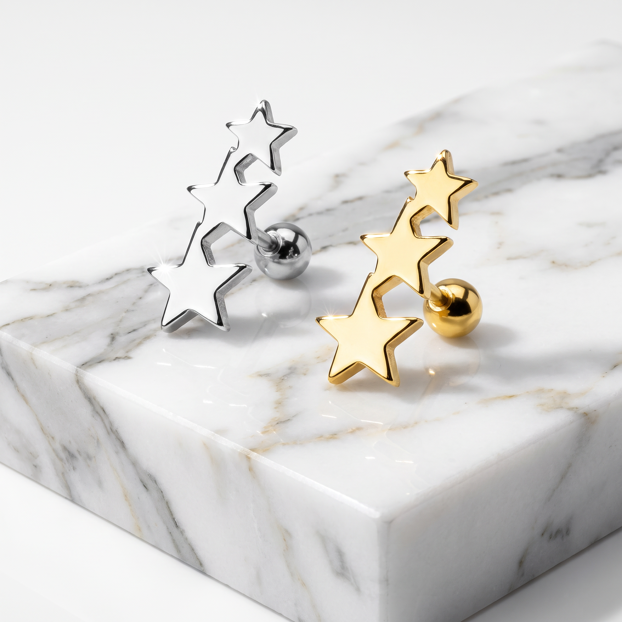 Two pairs of star-shaped stud earrings, one silver-toned and one gold-toned, displayed on a white marble block against a clean white background.