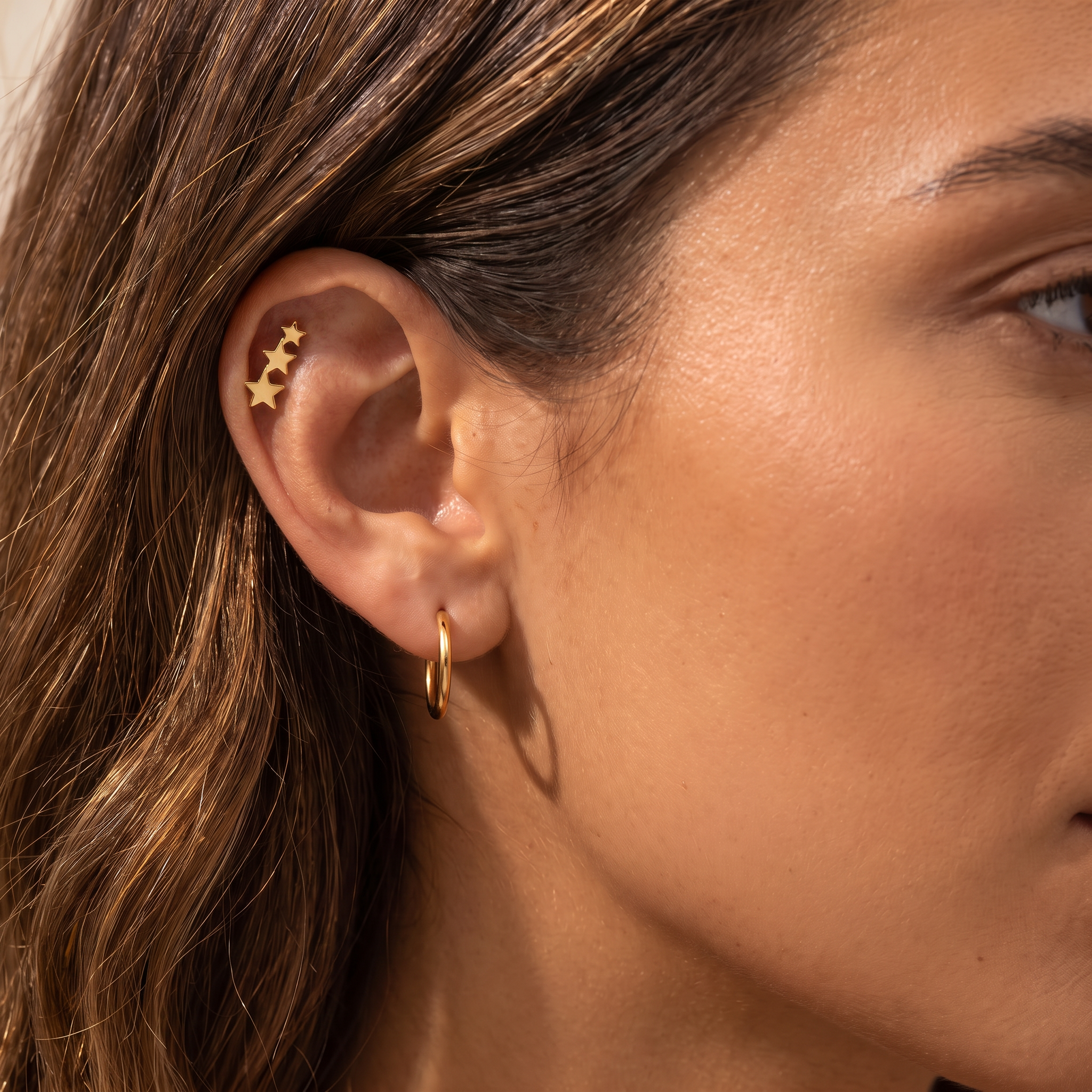 Close-up of a woman's ear showcasing a gold ear cuff with three stars and a small gold hoop earring.