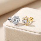 Two small flower-shaped stud earrings displayed on a cream jewelry box. One earring is silver-toned with a clear center stone and small clear stones around it, and the other is gold-toned with the same design.