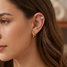 A close-up of a woman's ear wearing a small gold hoop earring and a gold ear cuff with a clear gemstone cluster, shown against her hair and skin.