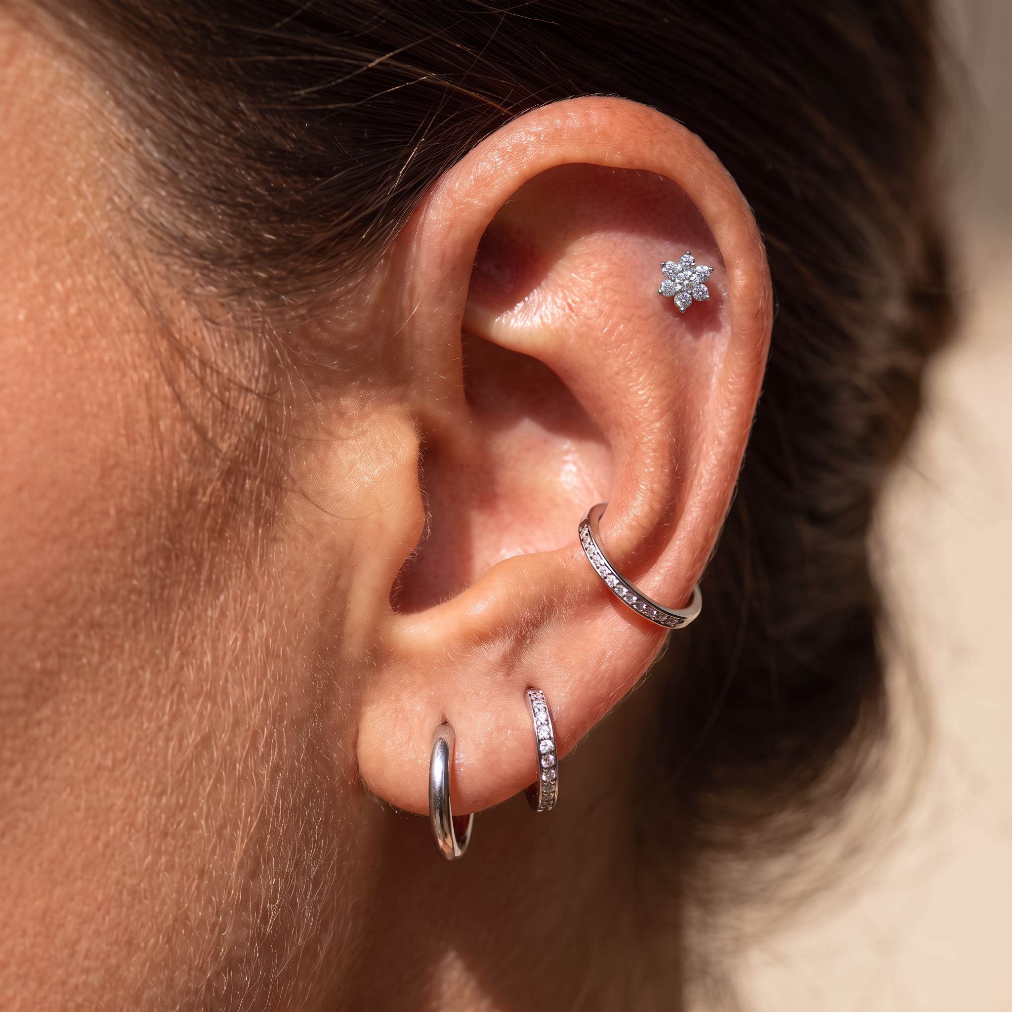 Close-up of a person's ear showcasing multiple silver piercings: a small flower-shaped stud in the lobe, a curved hoop in the helix, a small vertical hoop in the ear cartilage, and a thin vertical band with tiny clear stones in the lobe.