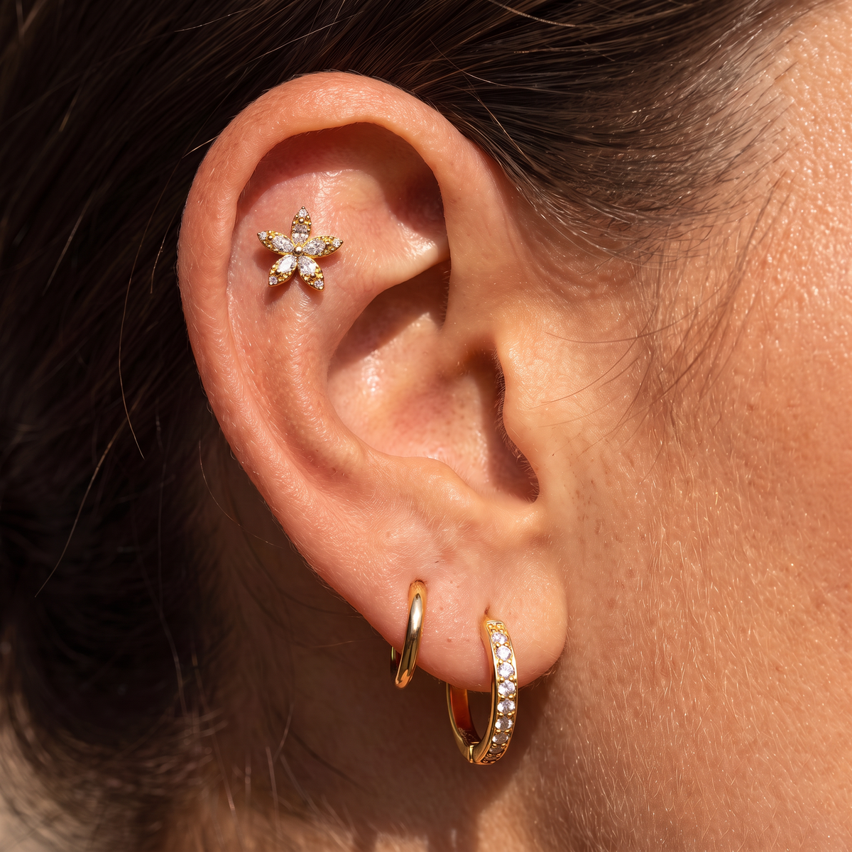 Close-up of a person's ear showcasing multiple gold ear piercings: a small gold flower stud on the upper ear cartilage, a gold hoop in the lobe, and a gold huggie hoop paved with clear crystals.