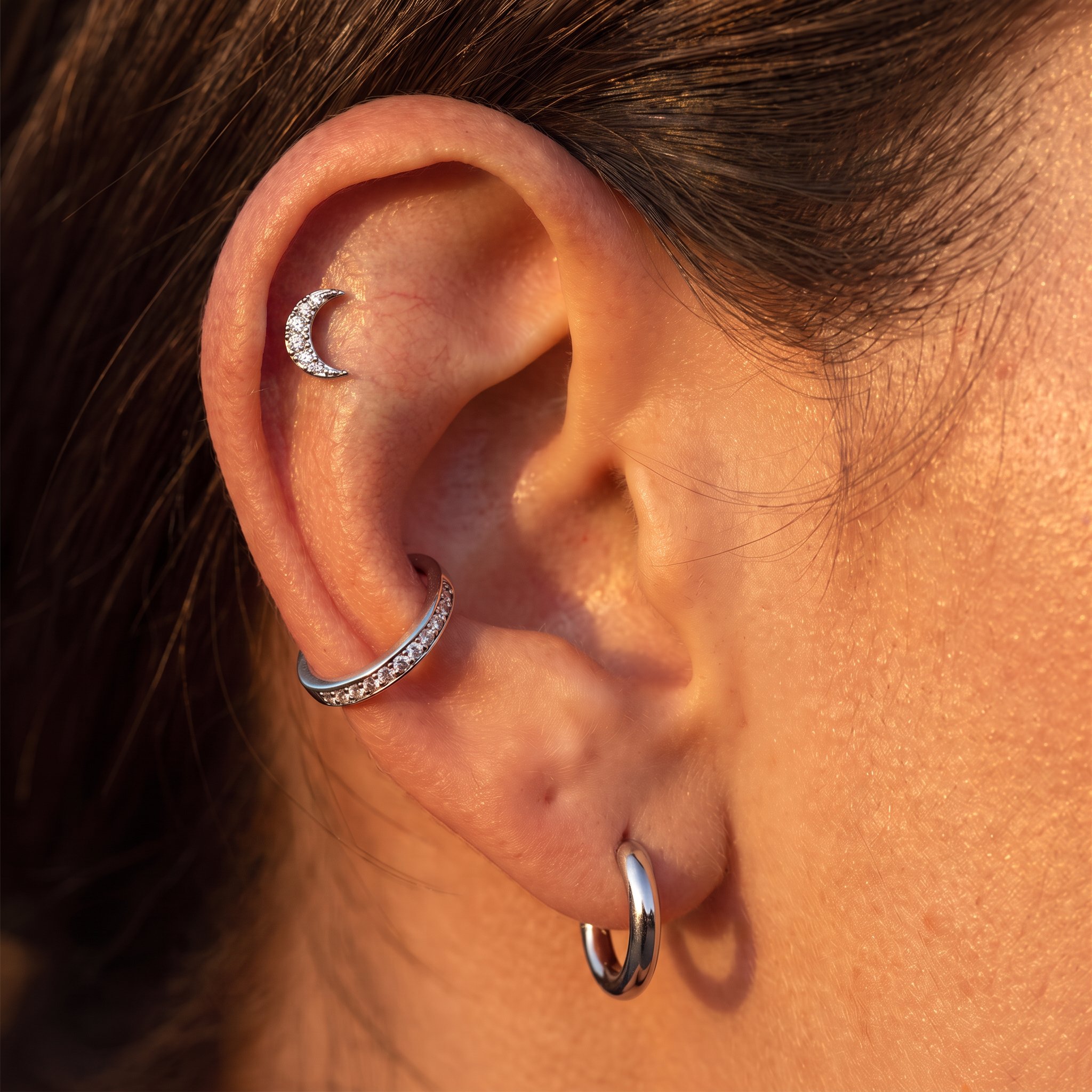 Close-up of a person's ear showcasing three silver ear piercings: a crescent moon stud in the cartilage, a curved hoop in the lobe, and a small silver hoop in the lobe. The warm golden-hour lighting highlights the skin and metallic shine of the jewelry.