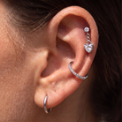 A close-up of a person's ear showcasing a multi-piece silver ear piercing set, including a hoop in the lobe, a curved cartilage hoop with pavé stones, and a small dangling heart-shaped cartilage stud.