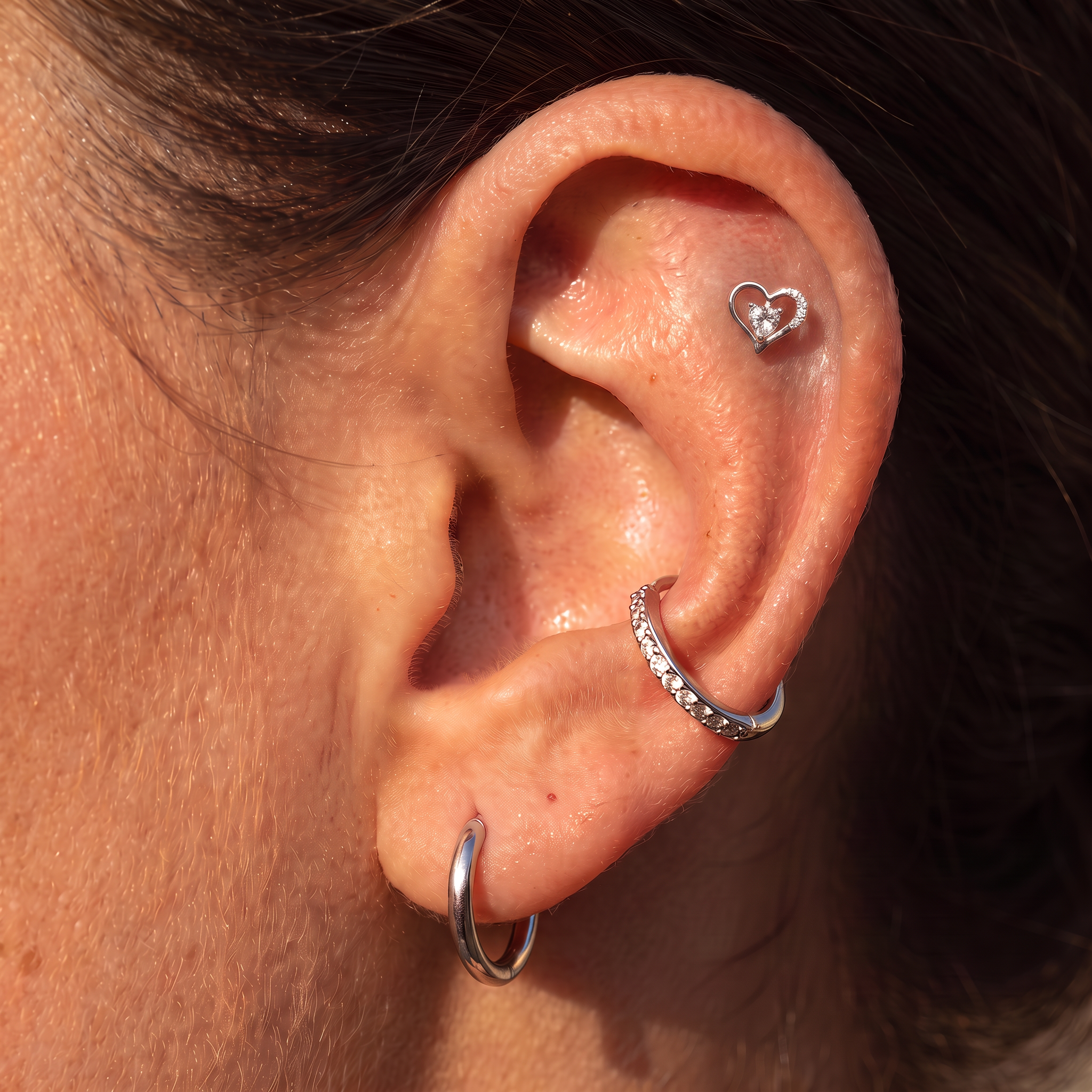 A close-up of a person's ear showcasing a four-hole ear cuff stack: a small silver heart stud in the lobe, a curved silver hoop with clear stones, a silver helix hoop, and a flat silver cartilage hoop.