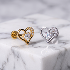 Two heart-shaped stud earrings displayed on a white marble block. One earring is gold-toned with a clear heart-shaped gemstone and small clear stones along the upper curve.