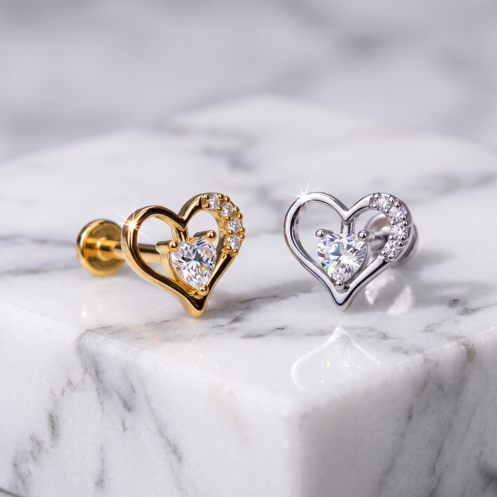 Two heart-shaped stud earrings displayed on a white marble block. One earring is gold-toned with a clear heart-shaped gemstone and small clear stones along the upper curve.