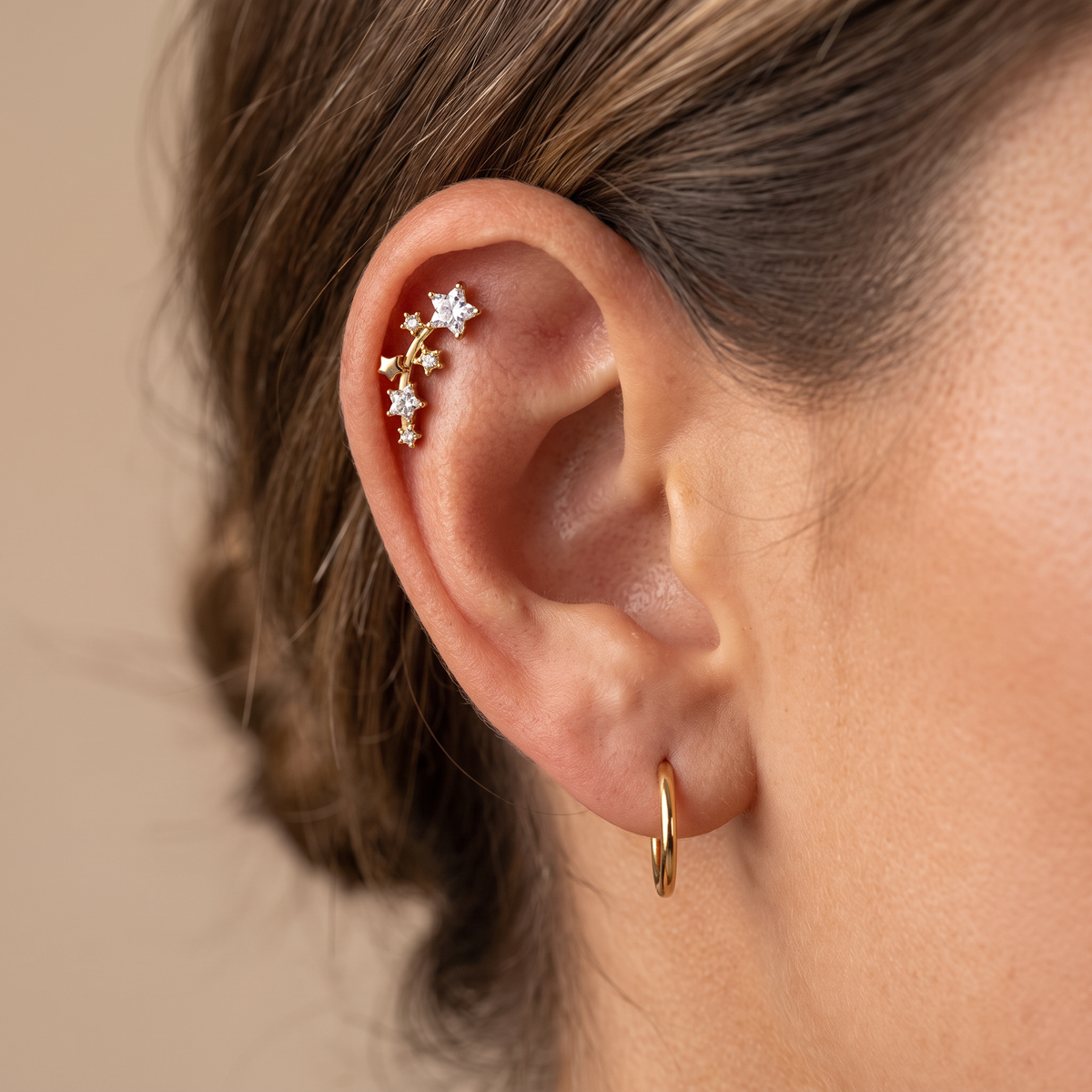 A close-up of a model's ear showcasing a gold helix stud adorned with small star-shaped crystal studs, paired with a simple gold hoop earring.