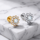 Two flower-shaped stud earrings with clear gemstones, one in yellow gold and one in silver, displayed on a white marble surface.