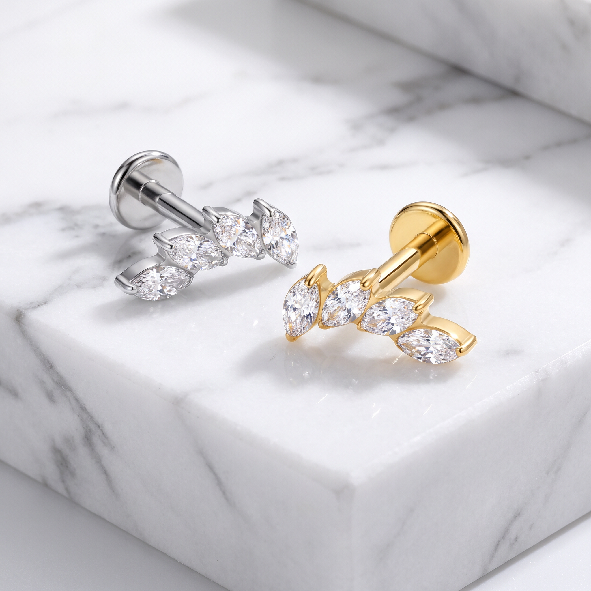 Two small stud earrings displayed on a white marble slab. One earring is silver-toned with marquise-cut clear stones, and the other is gold-toned with matching clear stones.