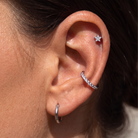 Close-up of a person's ear showcasing three silver ear piercings: a small star-shaped stud in the upper cartilage, a curved hoop lined with clear stones along the ear canal, and a simple silver hoop in the lobe.